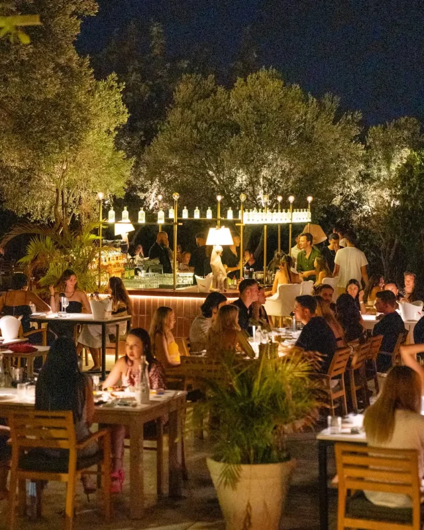 Alaçatı'nın en iyi mekanları arasında yer alan Yeniyer'in ağaçlarla çevrili açık hava restoranında, misafirlerin uzun yaz akşamlarında yemek ve kokteyl eşliğinde keyifli vakit geçirdiği canlı atmosfer.
