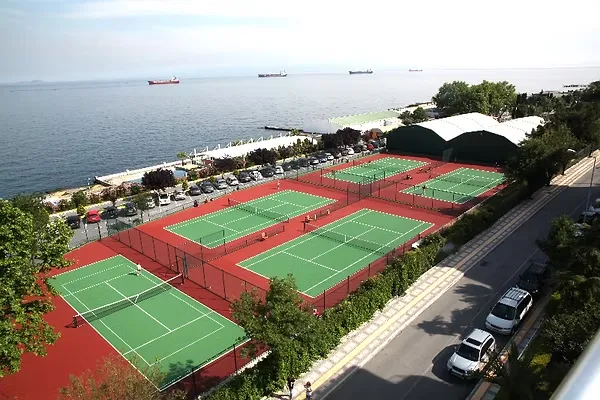 Yeşilyurt Tenis Spor Kulübü'nün deniz kenarında konumlanmış, yemyeşil kortları ve kırmızı kaplamalı zeminleriyle dikkat çeken birden fazla açık hava tenis kortunu, arka planda gemilerin yüzdüğü geniş denizi ve kulüp binalarını içeren genel bir görünüm.