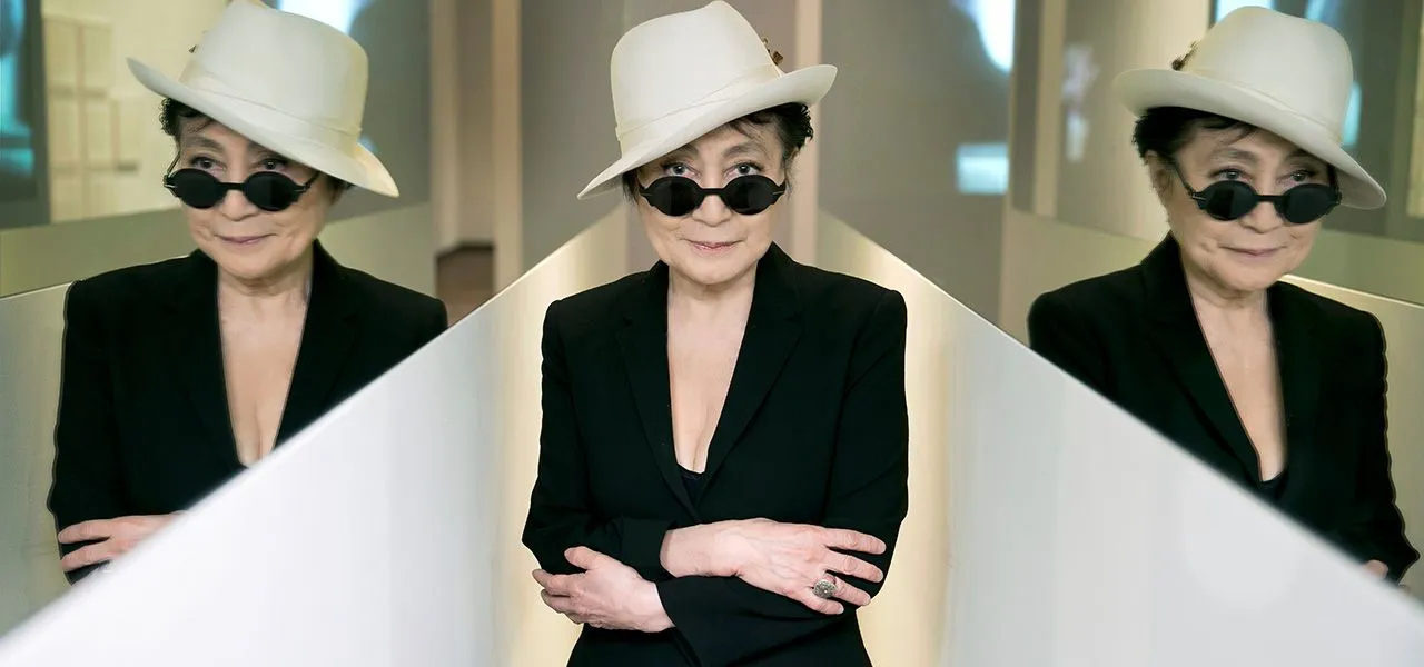 Krem rengi şapkası, küçük yuvarlak siyah güneş gözlükleri ve siyah blazer ceketiyle, hafifçe gülümseyerek doğrudan objektife bakan Yoko Ono, Sakıp Sabancı Müzesi'nde açılacak Yoko Ono: Insound and Instructure sergisi için çekilmiş bu fotoğrafta, arka plandaki yansımalarla çoğaltılmış görüntüsüyle çağdaş sanatın zamansız bir figürü olarak duruyor.