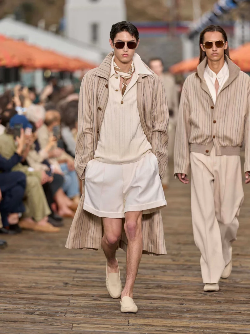 Zegna İlkbahar-Yaz 2027 defilesinden, Malibu sahilinde rahat terzilik anlayışını yansıtan, çizgili bej trençkot, açık renk polo yaka tişört, beyaz şort ve hasır ayakkabılar giyen güneş gözlüklü bir erkek modelin akışkan adımlarla yürüdüğü, arkasında da benzer hafif tonlarda bir diğer modelin göründüğü bir an.