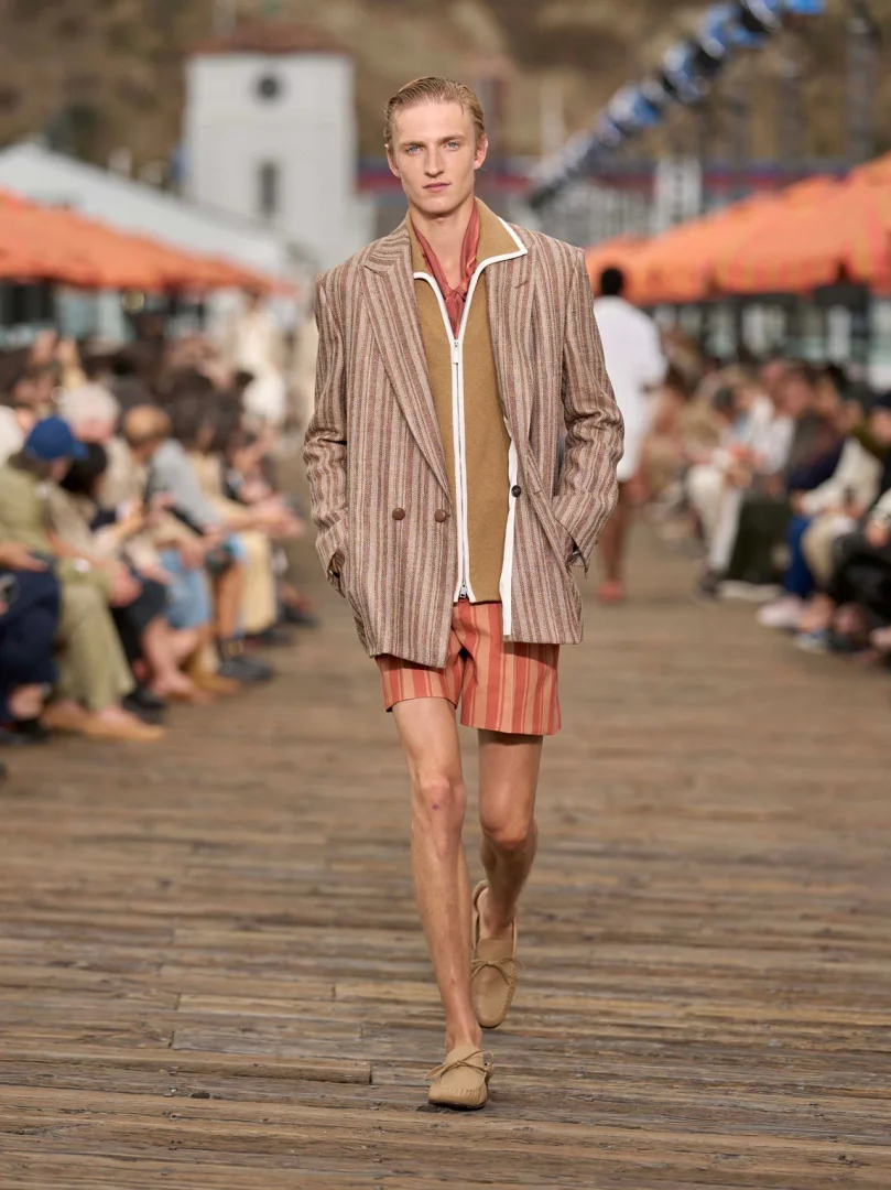 Malibu Pier'de yürüyen model, Zegna İlkbahar-Yaz 2027 erkek modası koleksiyonundan kahverengi-bej çizgili blazer ceketi, ona uyumlu şortu ve hafif mokasenleriyle yaşanmış lüks ve rahat terzilik anlayışını yansıtan şık bir yaz görünümü sergiliyor.