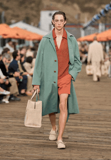 Zegna İlkbahar-Yaz 2027 erkek defilesi için Malibu sahildeki ahşap podyumda yürüyen bir model, mint yeşili bol bir trençkotun altında pas rengi V yaka örgü üst ve şort takımıyla, bej bağcıklı loafer ayakkabıları ve elinde açık renk bir bez çantasıyla akışkan terzilik ve rahat şıklığı sergiliyor.