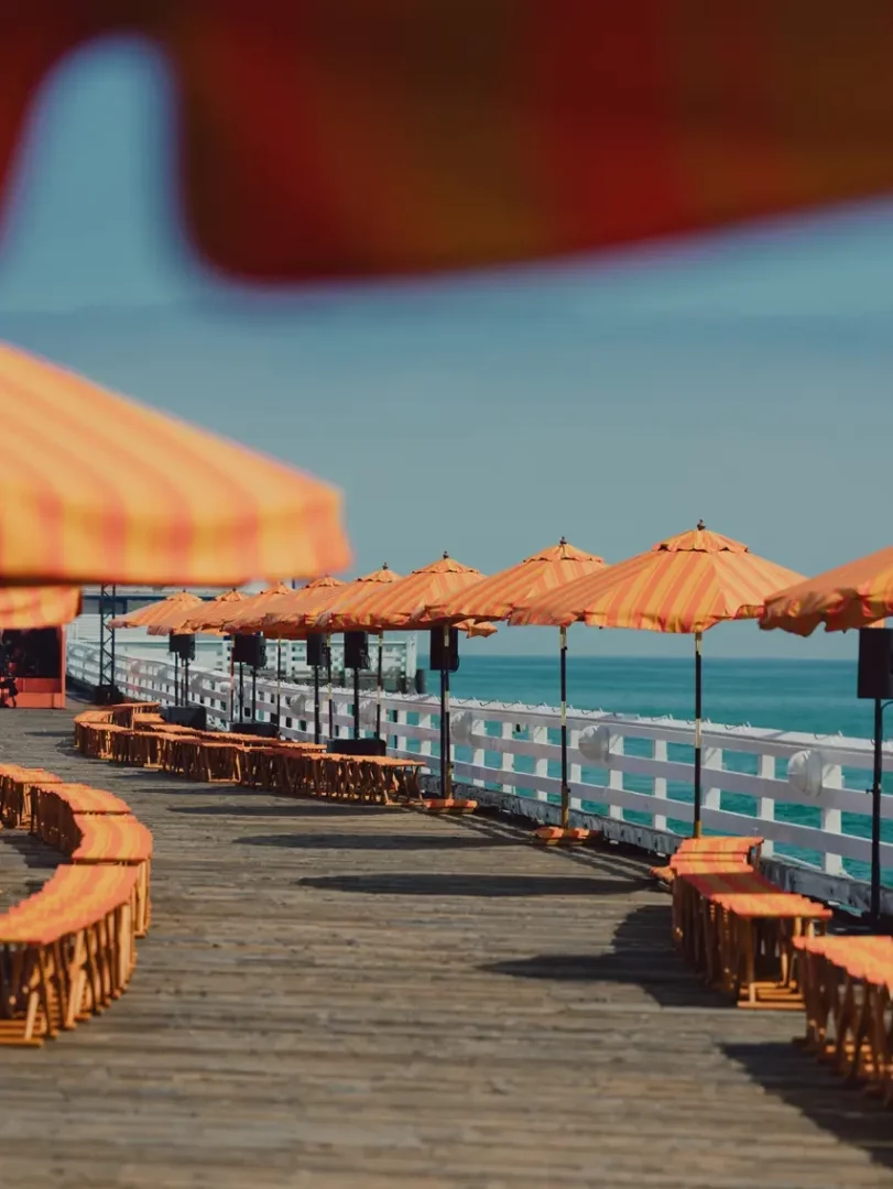 Malibu iskelesinde yer alan, turuncu ve krem rengi şeritli şemsiyelerle gölgelenmiş, uzun ahşap bir yürüyüş yolunun masmavi Pasifik Okyanusu'na doğru uzandığı Zegna İlkbahar-Yaz 2027 erkek giyim defilesi alanı, İtalyan yazının kaygısız ruhunu taşıyan rahat şıklığı vurguluyor.