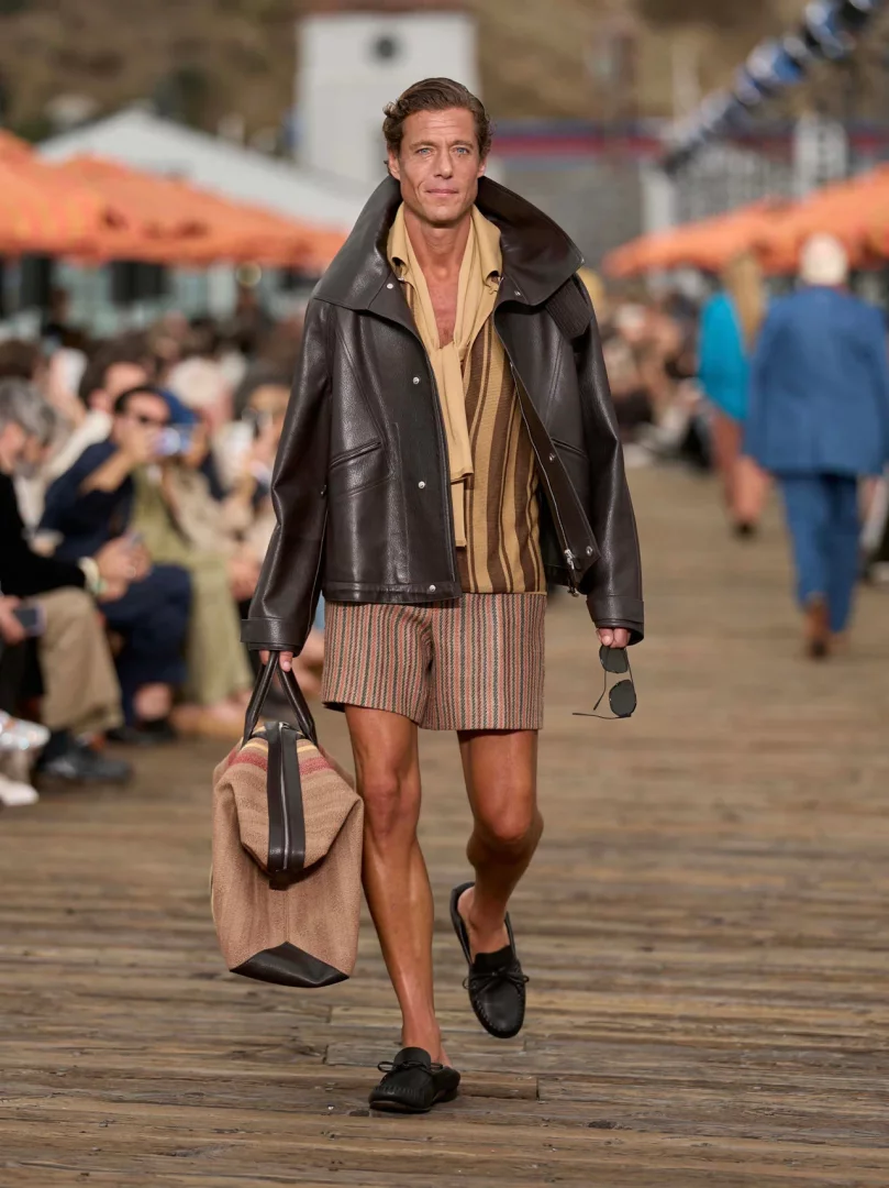 Alessandro Sartori'nin La Villeggiatura yaklaşımını Malibu defilesine taşıyan Zegna İlkbahar-Yaz 2027 erkek giyim koleksiyonundan bir model, açık renk çizgili şortu, rahat kesim kahverengi deri ceketi ve omuz askılı çantasıyla, akışkan terzilik ve kaygısız bir şıklık sunan stilini podyumda sergiliyor.