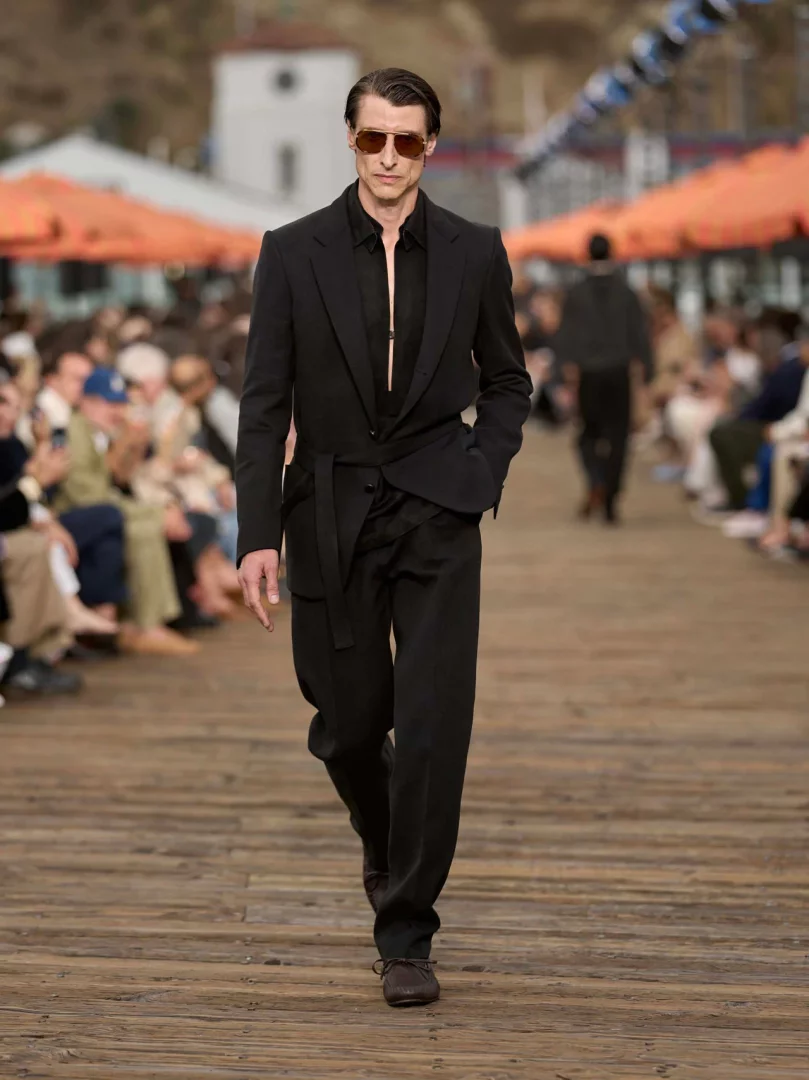 Siyah, bağlamalı kemerli, rahat kesim bir takım elbise ve açık yakalı bir gömlek giymiş, güneş gözlüklü bir erkek model, Malibu İskelesi'ndeki Zegna İlkbahar-Yaz 2027 erkek giyim defilesinin ahşap podyumunda kendinden emin adımlarla yürüyor, koleksiyonun yaşanmış lüks ve akışkan terzilik anlayışını yansıtıyor.
