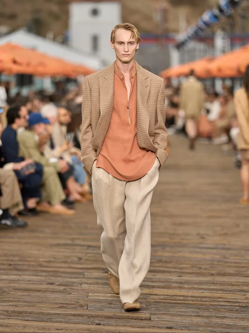 Malibu Pier'de düzenlenen Zegna İlkbahar-Yaz 2027 erkek giyim defilesinden bir an: Bol kesimli, doğal tonlarda bej keten pantolon, yıkanmış görünümlü şeftali rengi bir gömlek ve hafif dokulu kahverengi blazer ile podyumda yürüyen model, La Villeggiatura felsefesinin getirdiği rahat ve akışkan terzilik anlayışını yansıtıyor.