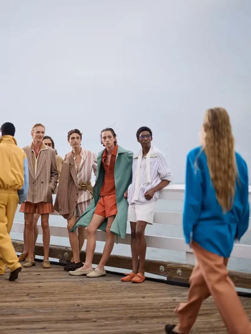 Zegna İlkbahar-Yaz 2027 erkek giyim koleksiyonundan, Malibu iskelesinde okyanus manzarası önünde duran, bej, sarı, pas ve nane yeşili tonlarındaki rahat kesim, çizgili ve düz ceketler, gömlekler ve şortlarla modern ve kaygısız bir yaz stili sunan modeller.