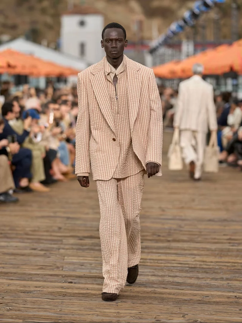 Malibu Pier'de düzenlenen Zegna İlkbahar-Yaz 2027 defilesinde, akışkan terzilik anlayışını yansıtan, bej rengi dikey çizgili ve belirgin dokulu hafif bir takım giyen, koyu tenli erkek model podyumda yürüyor.