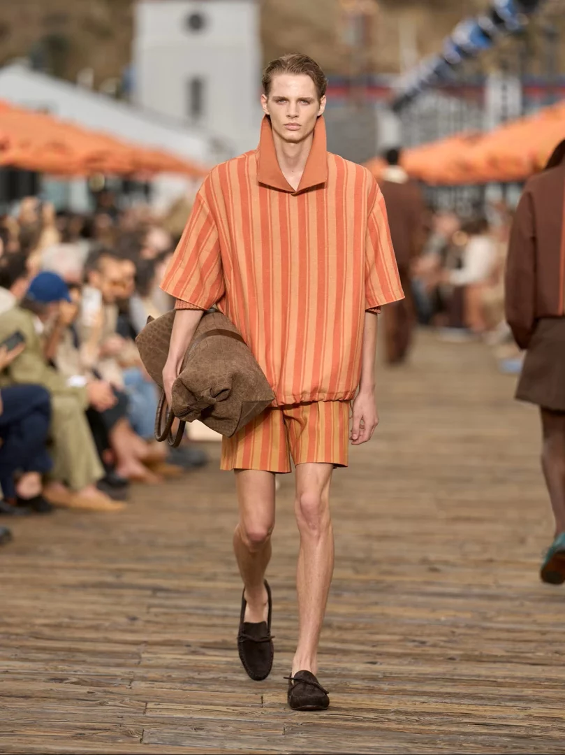 Malibu Pier'de yürüyen bir erkek model, Zegna İlkbahar-Yaz 2027 koleksiyonundan turuncu ve bej dikey çizgili, gevşek kesimli, polo yaka kısa kollu bir üst ve uyumlu şort giyiyor, sol kolunda süet görünümlü kahverengi bir seyahat çantası taşıyarak La Villeggiatura felsefesinin rahat ve yaşanmış lüks stilini öne çıkarıyor.