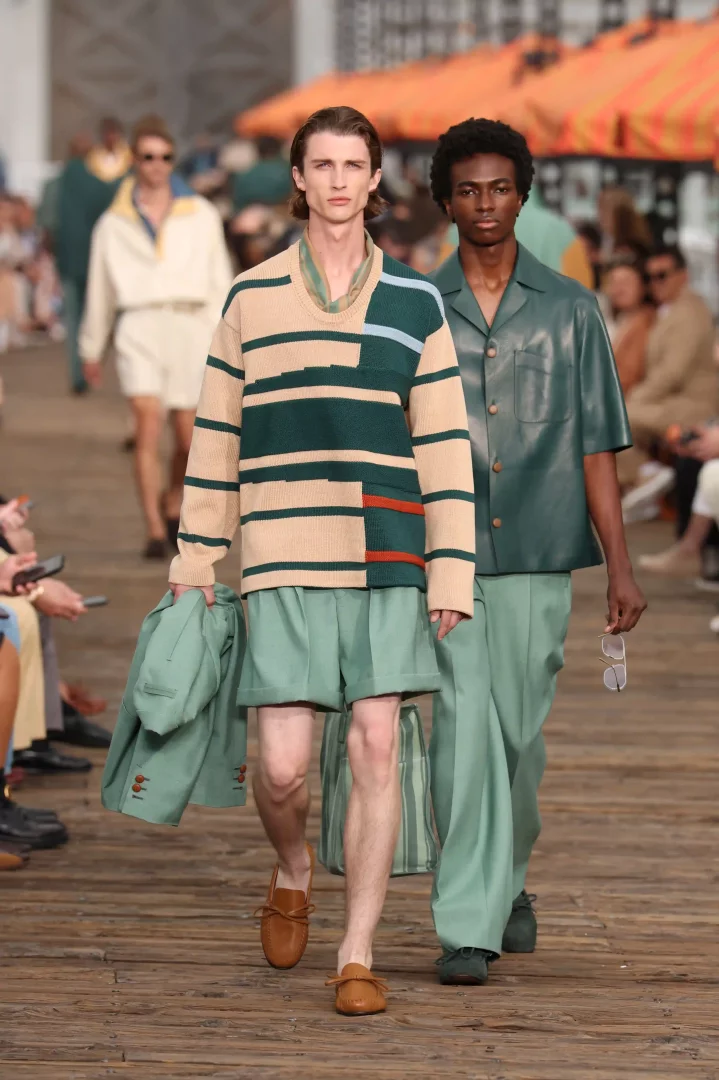 Zegna İlkbahar-Yaz 2027 erkek giyim koleksiyonundan iki model, bej ve nane yeşili tonlarındaki rahat çizgili triko kazaklı ve şortlu ilk model ile koyu nane yeşili akışkan gömlek-pantolon takımı giyen ikinci modelle podyumda ilerleyerek La Villeggiatura ruhunu yansıtan modern ve kaygısız bir yaz şıklığı sunuyor.