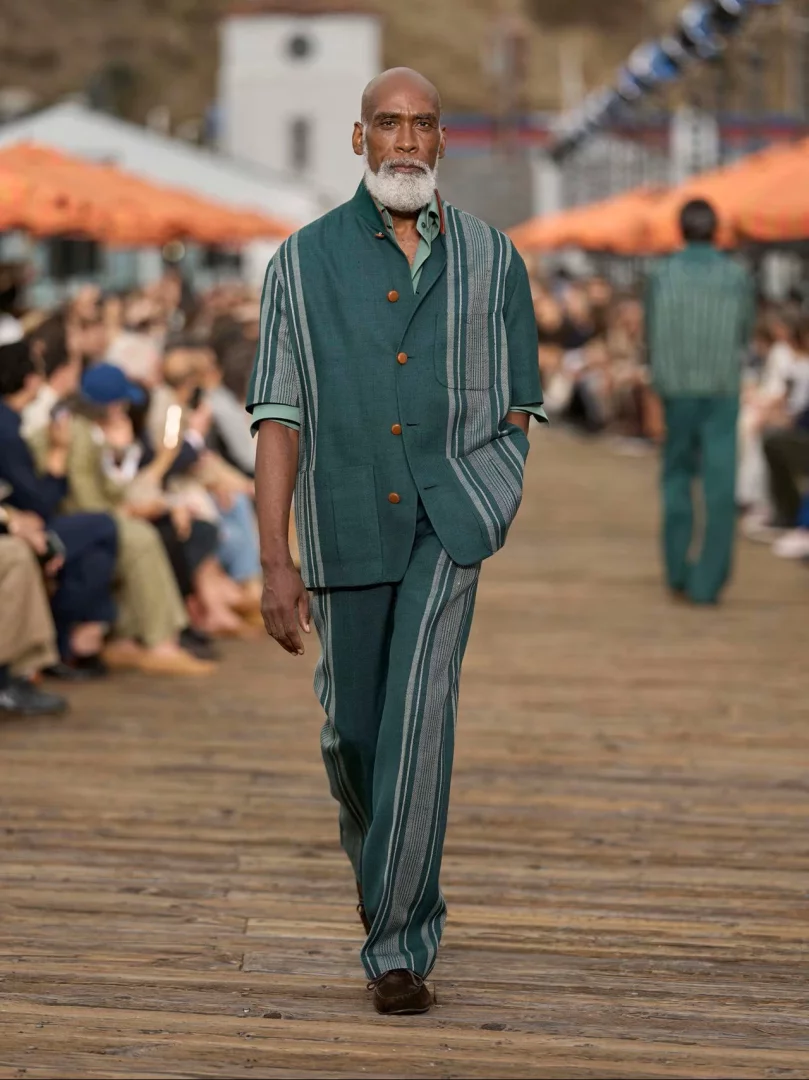 Zegna İlkbahar-Yaz 2027 erkek giyim koleksiyonundan, Malibu İskelesi'nde ahşap bir podyumda ilerleyen beyaz sakallı bir model, yanlarında açık gri dikey şerit desenleri ve kahverengi düğmeleri olan petrol yeşili, kısa kollu bir ceket ile dış kenarları çizgili, bol kesimli uyumlu pantolonu ve açık yeşil gömleğiyle koleksiyonun akışkan terzilik ve rahat şıklık anlayışını yansıtıyor.