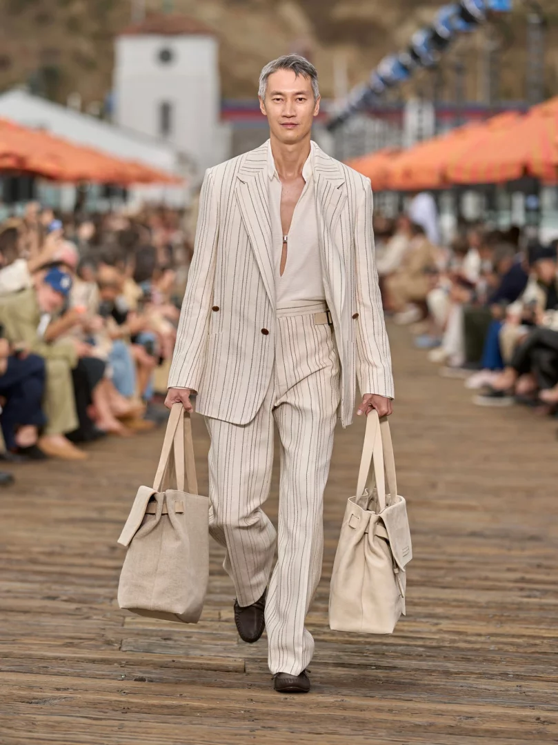 Malibu İskelesi'nde gerçekleştirilen Zegna İlkbahar-Yaz 2027 erkek giyim defilesinde, çizgili, bej bir takım elbise giymiş ve iki elinde keten görünümlü çanta taşıyan bir model, koleksiyonun rahat terzilik ve yaşanmış lüks anlayışını yansıtan akışkan ve hafif bir duruşla yürüyor.