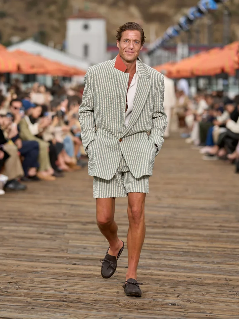 Model, Malibu İskelesi'nde düzenlenen Zegna İlkbahar-Yaz 2027 defilesinde, hafif ve akışkan terzilik anlayışını yansıtan, çizgili, açık yeşil şortlu takım elbisesi ve rahat mokasenleriyle özgür ruhlu bir yaz şıklığı sergiliyor.