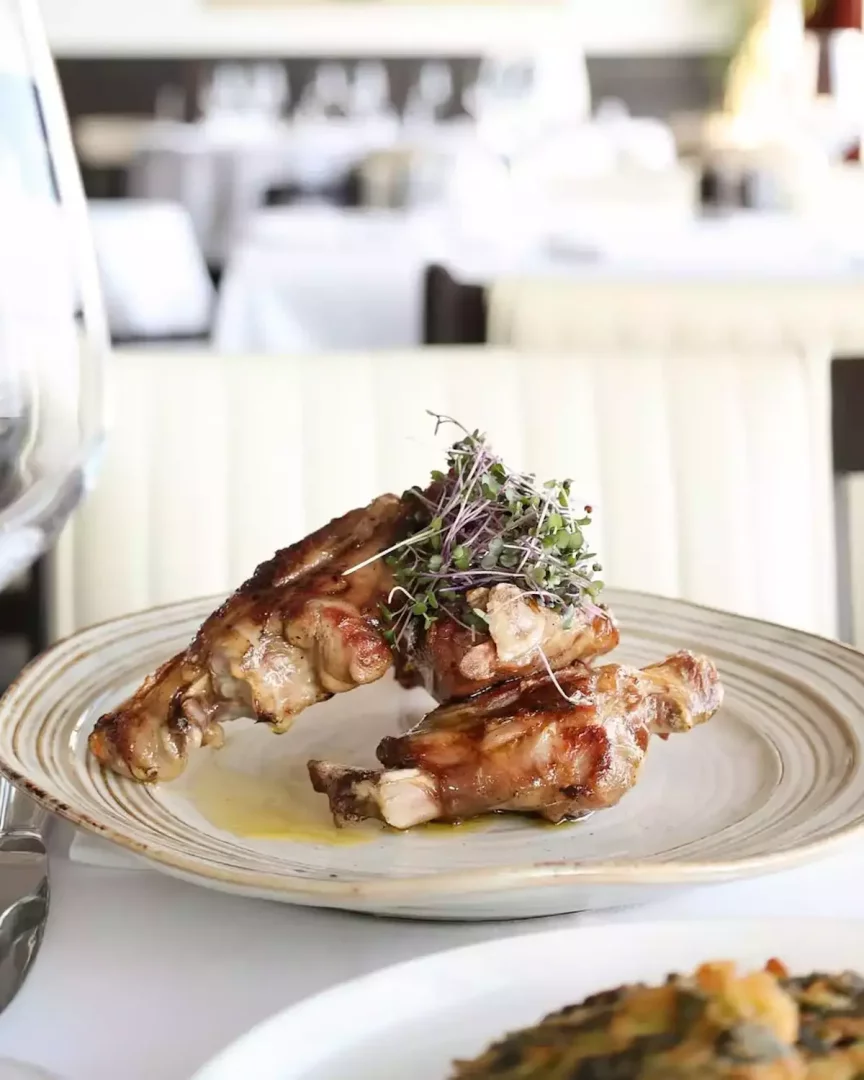 Antalya'nın meşhur 7 Mehmet restoranında servis edilen, nar gibi kızarmış üç parça kuzu kaburga, mor ve yeşil filizlerle süslenmiş, kenarları desenli açık renk bir tabağın üzerinde iştah açıcı bir şekilde duruyor; arka planda lüks bir restoranın beyaz masa örtüleri ve loş ortamı seçiliyor.
