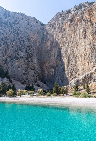 Simi Adası'nda yer alan Agios Georgios plajında, kristal berraklığındaki turkuaz sular, beyaz çakıllı kıyı şeridi, sahildeki ağaçlar ve hemen arkasında yükselen heybetli, sarp kayalıkların çevrelediği küçük bir şapelin de görüldüğü muhteşem bir koy manzarası.