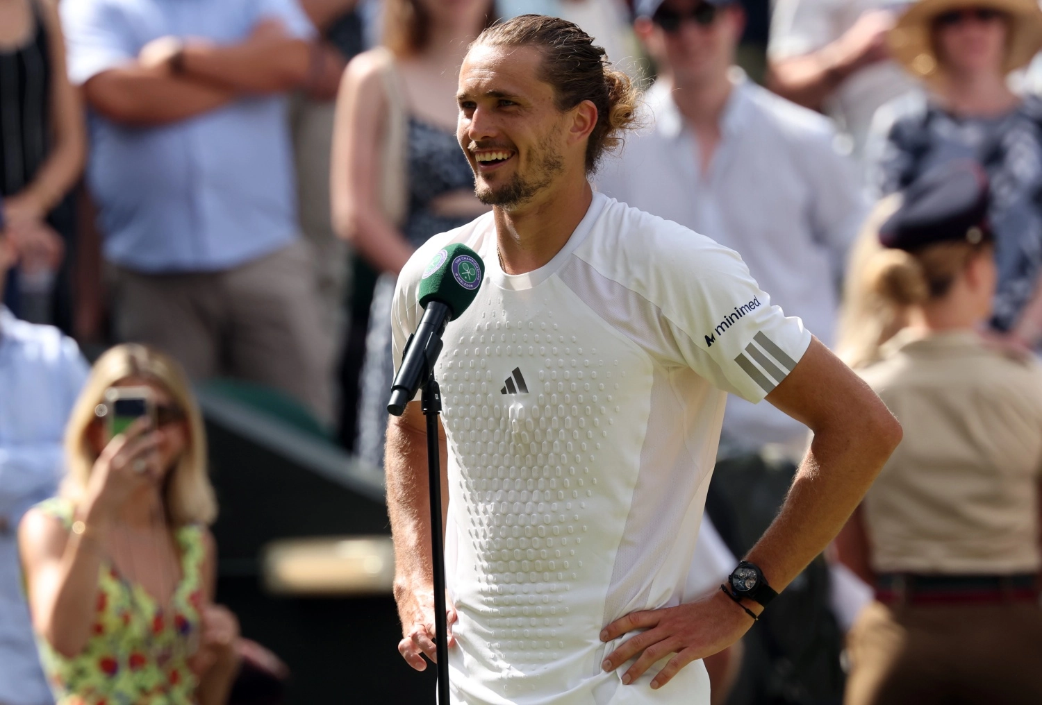 Alexander Zverev, Wimbledon logosu taşıyan bir mikrofonun önünde samimi bir gülümsemeyle poz verirken, üzerinde beyaz tenis tişörtü ve sol bileğinde Jacob & Co Epic X Black kol saati belirgin bir şekilde görülüyor.