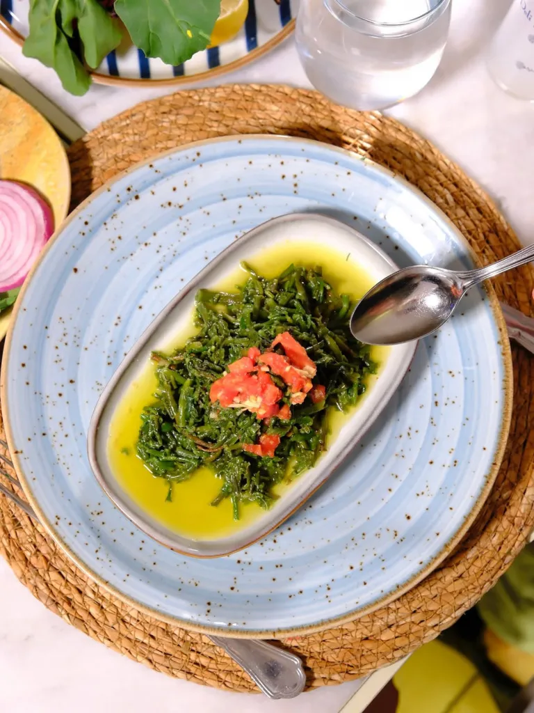 Deli Yengeç restoranında sunulan, hasır altlık üzerindeki benekli açık mavi seramik tabağın içinde zeytinyağında hazırlanmış, üzerinde parlak kırmızı domates küpleriyle süslü, doyurucu bir yeşil ot mezesi yemeği yakın çekim; sağda yemeğe uzanan bir kaşık ve arka planda bulanık görünen diğer detaylarla birlikte, Akdeniz mutfağının lezzetini yansıtan keyifli bir başlangıç.