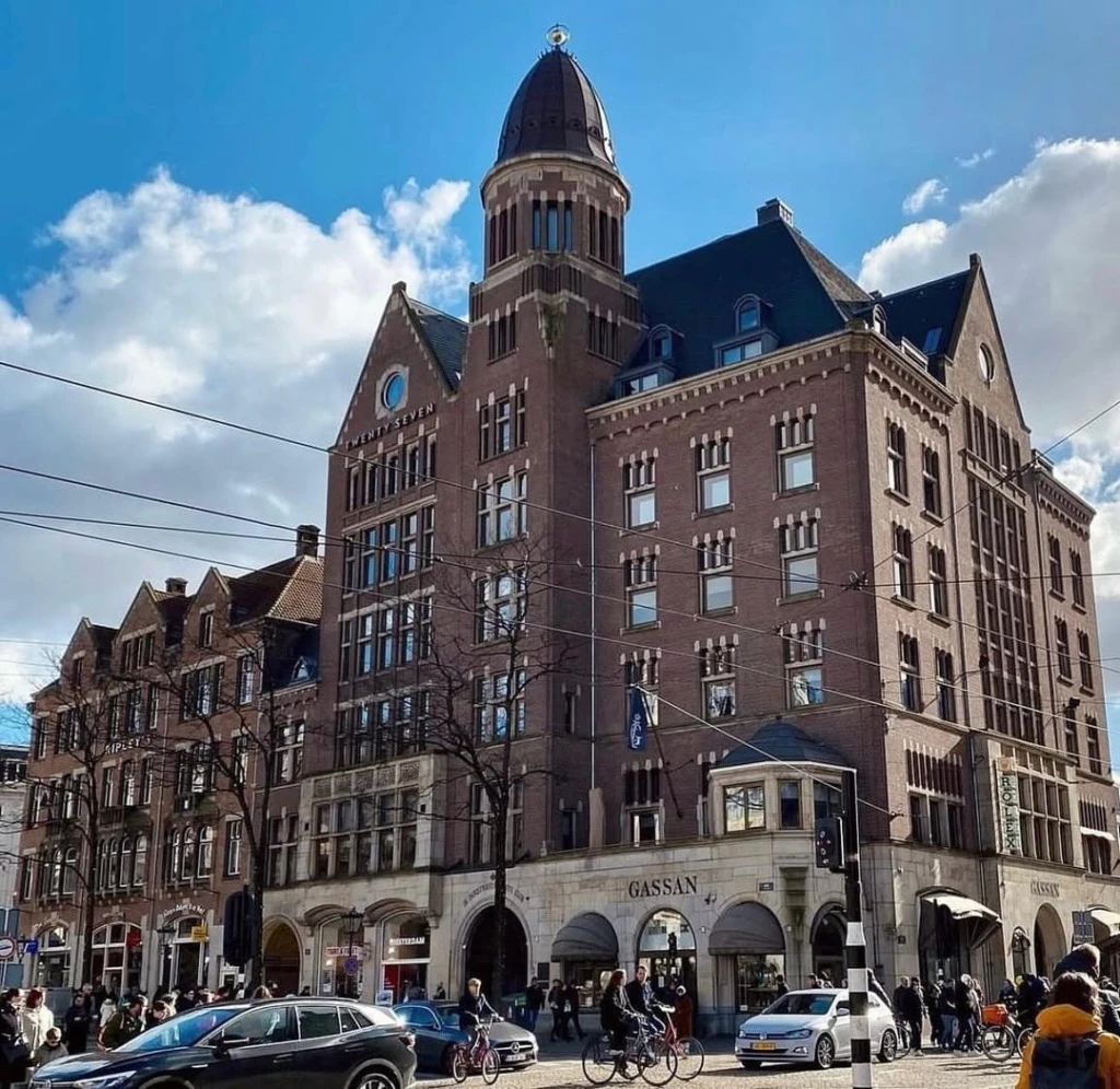 Amsterdam'ın hareketli merkezinde yer alan, çarpıcı kubbeli kulesi ve kırmızı tuğlalı dış cephesiyle dikkat çeken Hotel TwentySeven, önünden geçen araçlar ve yayalarla dolu bir caddede güneşli bir günde tüm ihtişamıyla yükseliyor.