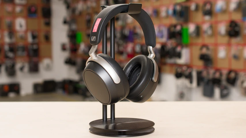Siyah ve gümüş renkli detaylara sahip, şık bir Sennheiser over-ear kulaklık, aydınlık bir masa üzerinde konumlanmış siyah bir kulaklık standında sergilenirken, arka planda bulanık bir elektronik ürün mağazası veya teşhir alanı seçiliyor.