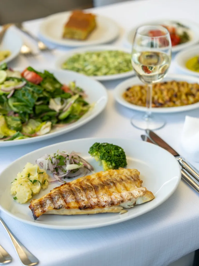 Sur Balık restoranında servis edilen ızgara balık filetosunun yanında püre, soğan salatası ve brokoliyle sunulduğu, masanın çeşitli taze mezeler ve bir kadeh beyaz şarapla zenginleştiği iştah açıcı bir yemek masası görüntüsü.