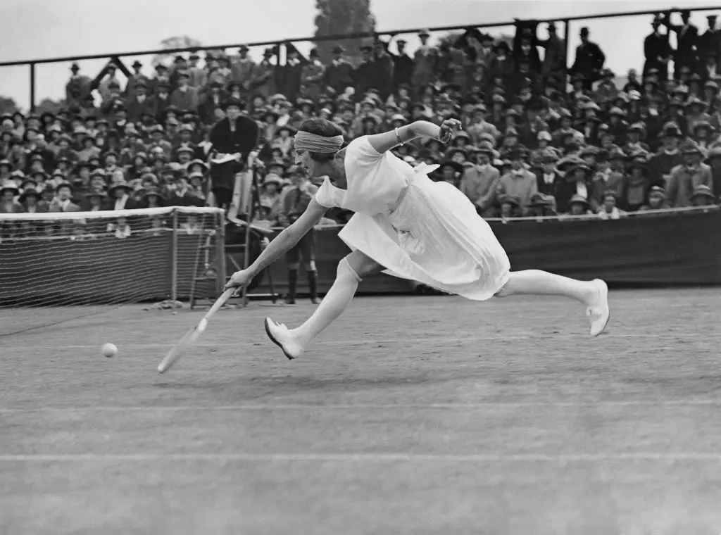 Kara beyaz fotoğrafta, beyaz elbise ve bandanayla Wimbledon kortunda tenis topuna raketle alçaktan uzanırken havada görünen efsanevi tenisçi Suzanne Lenglen, arkasında oturan seyircilerle birlikte dinamik bir anı yakalıyor.