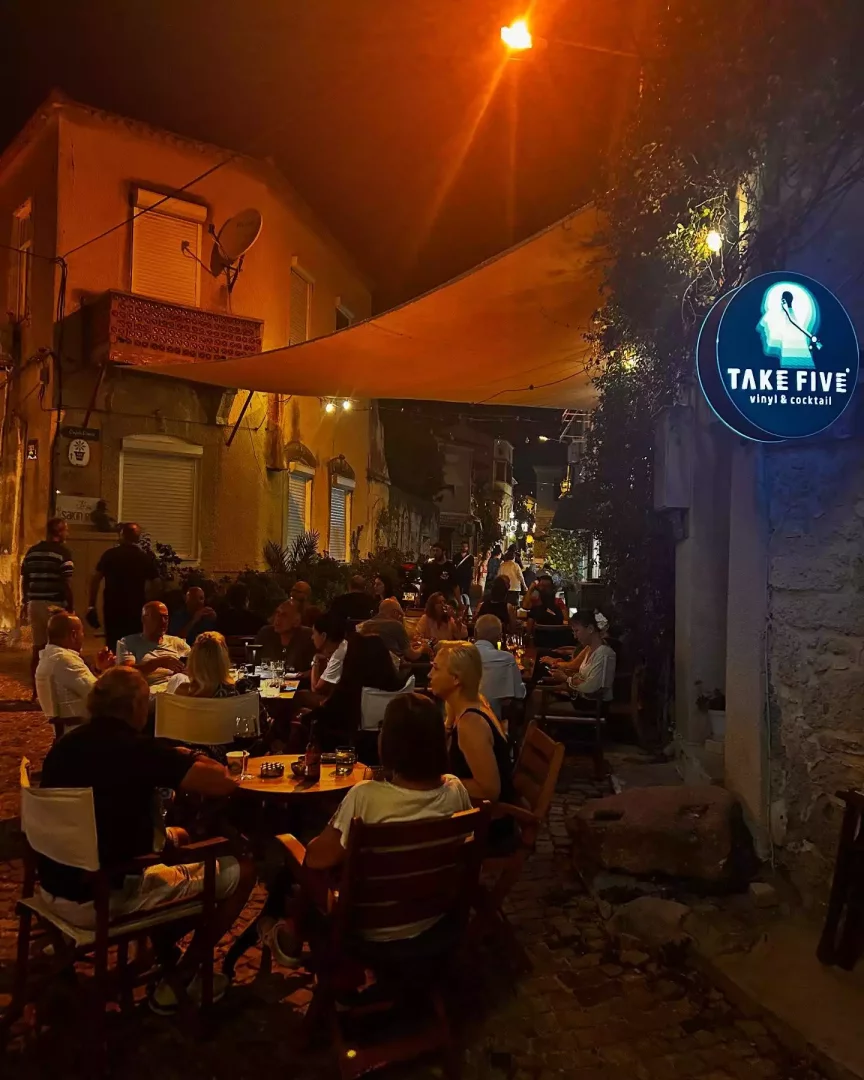 Turuncu sokak lambalarının ve gerilmiş tentenin aydınlattığı tarihi bir sokakta, Take Five vinyl & cocktail mekanının canlı dış oturma alanında, masalarda keyifli sohbetler eden ve içkilerini yudumlayan kalabalık bir grup insan görülüyor.