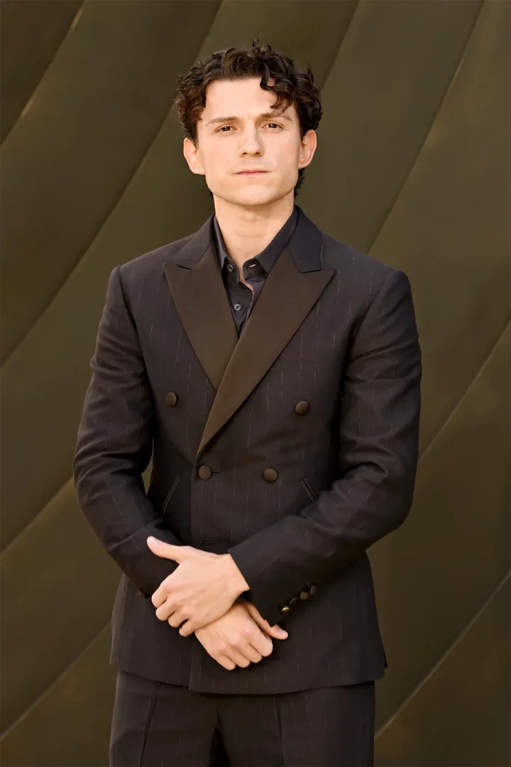 Tom Holland, koyu kıvırcık saçları, parlak saten yakalı ince çizgili koyu kruvaze takım elbisesiyle, ellerini önünde kavuşturmuş ciddi bir ifadeyle dururken The Odyssey filminin premiyer etkinliğinde çekilmiş bu fotoğrafta dikkat çekiyor.