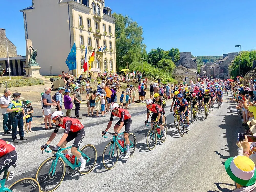 Tour de France 2025 Yedinci Etabı'nda, Corseul kasabasının güneşli bir gününde, tarihi bir binanın ve coşkulu seyircilerin önünden hızla geçen bisikletçiler pelotonu kıyasıya bir mücadele veriyor.