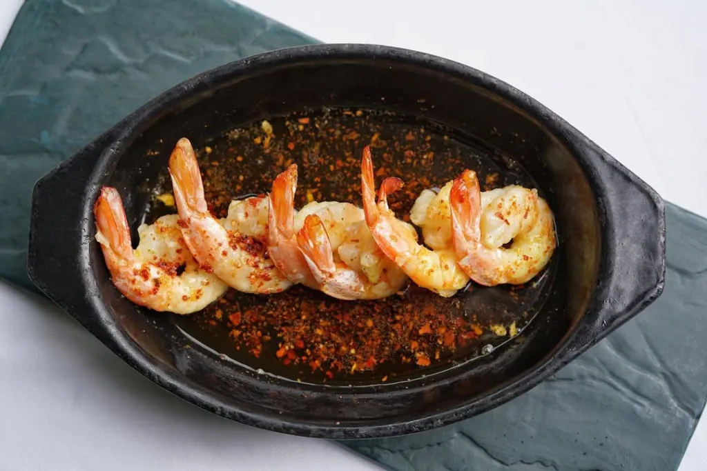 Trilye restoranında sunulan, koyu renkli, oval bir sahanın içinde bol zeytinyağı ve pul biber eşliğinde, kuyrukları yukarı doğru özenle dizilmiş altı adet parlak turuncu karidesin bulunduğu iştah açıcı bir deniz mahsulü tabağı koyu gri dokulu bir servis altlığı üzerinde.