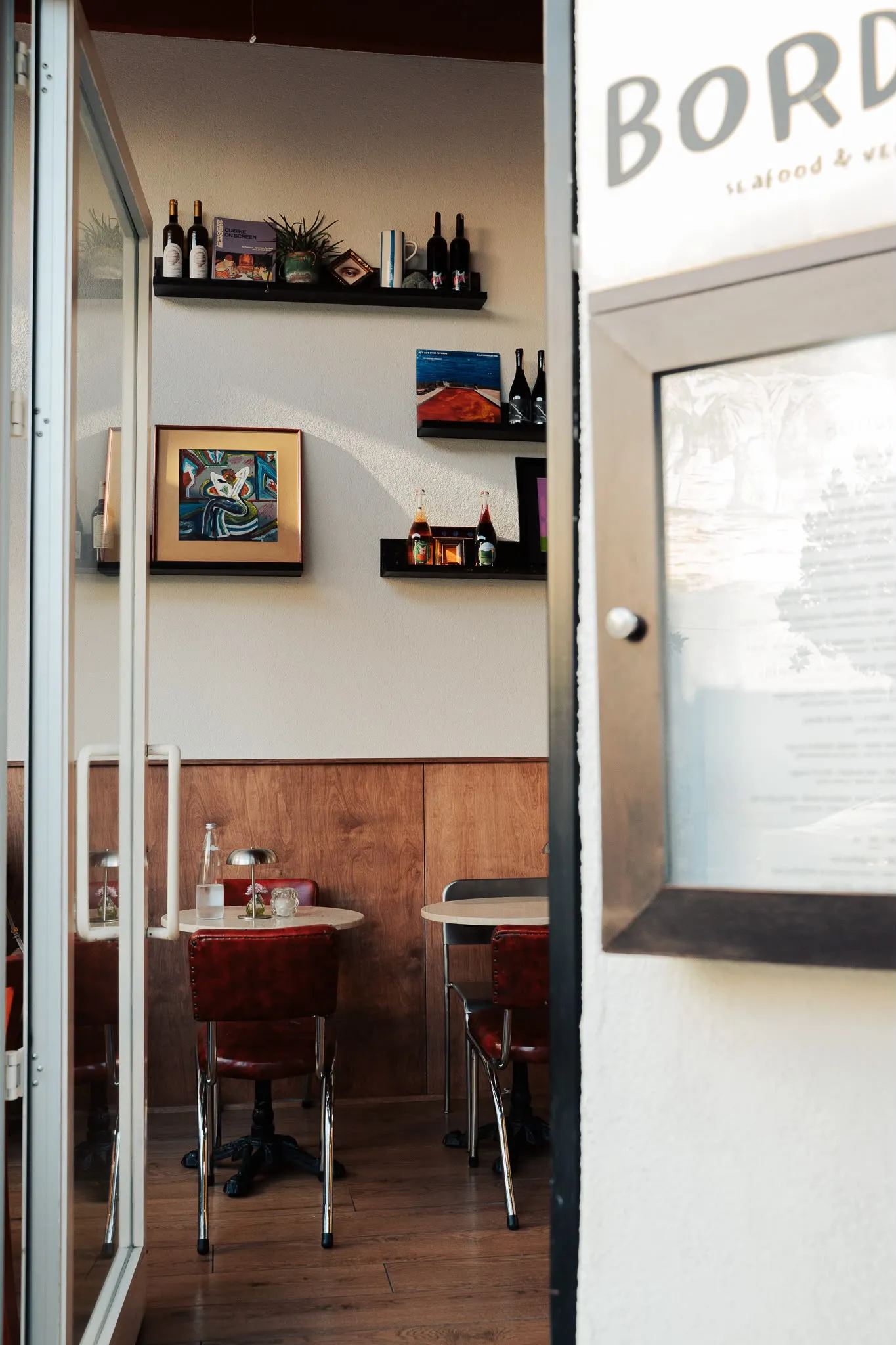 Açık cam kapıdan görünen, ahşap zemin ve panelli duvarlarla dekore edilmiş, güneş ışığı alan modern bir restoran iç mekanı; içerisinde küçük yuvarlak masalar, koyu kırmızı deri sandalyeler, duvardaki raflarda şarap şişeleri, kitaplar, bitkiler ve çerçeveli sanat eserleri ile sıcak ve davetkar bir yemek atmosferi sunuyor.