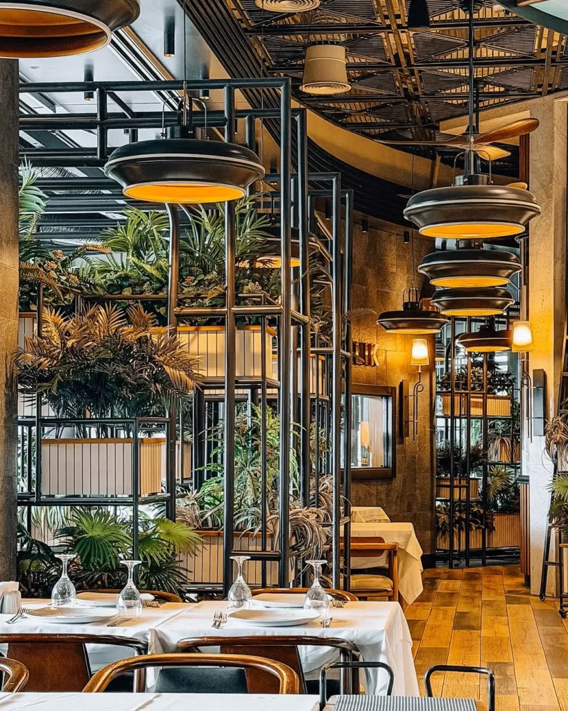 Yelken Balık restoranının şık iç mekanında, beyaz örtülü masalar misafirleri beklerken, modern siyah avizelerden yayılan sıcak ışıklar ve yeşil bitkilerle dolu dekoratif metal seperatörler çağdaş ve doğal bir ambiyans sunuyor.