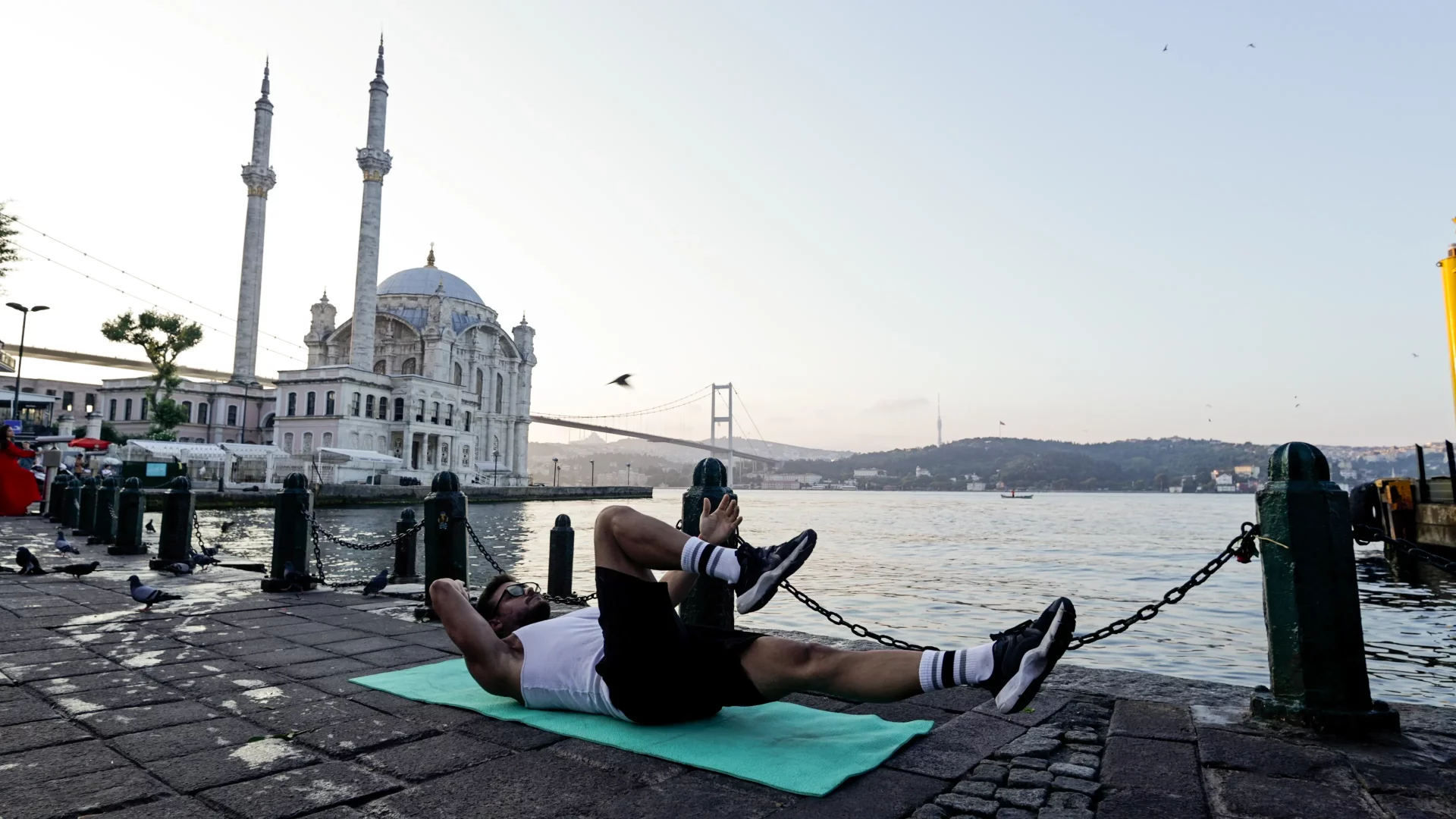 Ortaköy Camii'nin zarif mimarisi ve Boğaz Köprüsü'nün silueti eşliğinde, beyaz atletli ve siyah şortlu bir adam, yeşil bir mat üzerinde sırtüstü uzanmış, bacaklarını havaya kaldırarak aktif bir açık hava kardiyo egzersizi yapıyor.