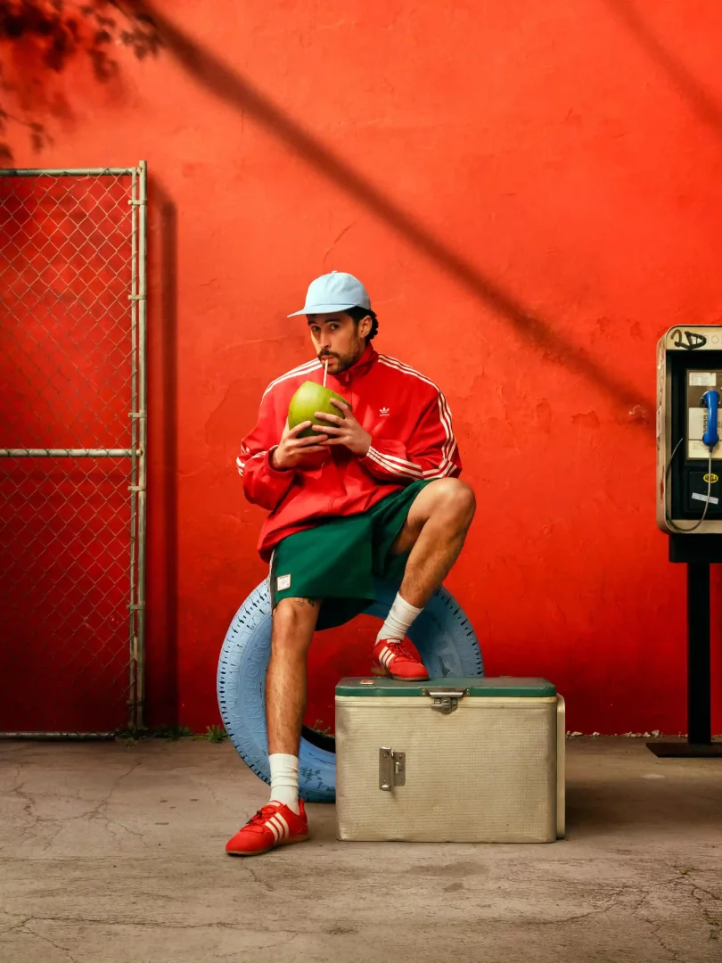 Bad Bunny, parlak kırmızı bir duvarın önünde, yeşil şortu, kırmızı Adidas ceketi ve ikonik kırmızı Adidas Samba ayakkabılarıyla mavi bir lastiğin ve bir soğutucunun üzerine oturmuş, elindeki taze hindistan cevizini yudumlarken görülüyor.