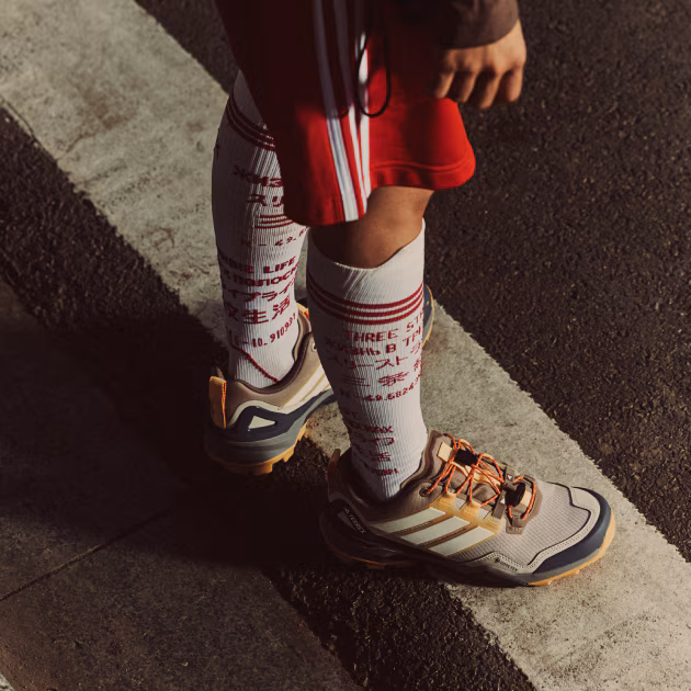 Kırmızı şort ve üzerinde çeşitli dillerde yazılar olan beyaz uzun çoraplar giyen bir kişinin ayakları, asfalt üzerindeki beyaz bir çizgide dururken bej ve lacivert tonlarında adidas Terrex Skychaser arazi ayakkabılarını sergiliyor.