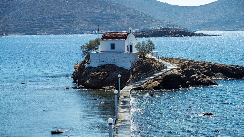 Denizin ortasında, kayalık bir adacığın üzerinde yer alan, beyaz badanalı, kırmızı kiremit çatılı şirin bir kilise görülüyor; Leros'taki bu ikonik Agios Isidoros yapısına uzanan, deniz yüzeyinde parlayan mavi suyun üzerinden geçen dar bir beton iskele ve basamaklar manzarayı tamamlıyor.