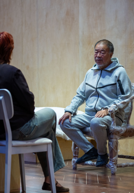 Manchester'daki Button Up! sergisinin bir parçası olan Sewing a Button performansı esnasında, sanatçı Ai Weiwei, gözaltı hücresinin ahşap duvarlı dar bir kopyasında gri eşofmanıyla otururken, kendisine gözetim ve iktidar temalı sorular yönelten bir gazeteciyle konuşuyor.
