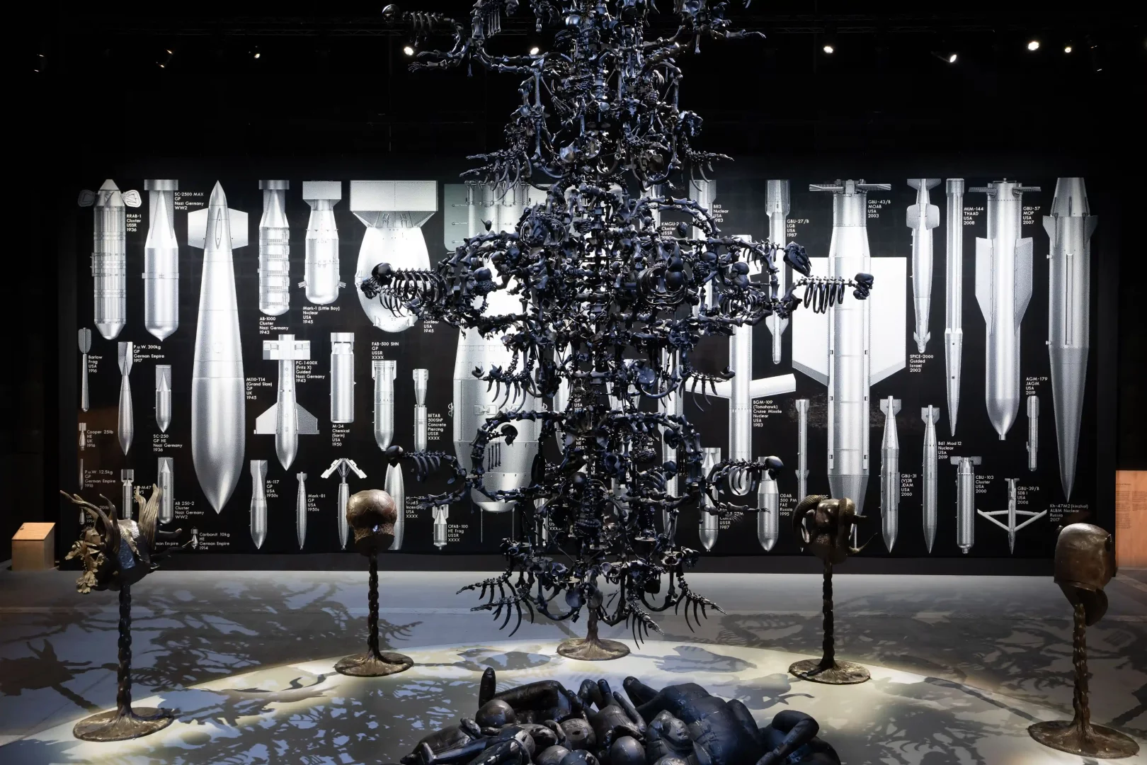 Ai Weiwei'nin Button Up! sergisindeki devasa, karanlık, iskeletvari ağaç enstalasyonu, arkasında sıralanmış farklı dönemlere ait bomba çizimleri içeren History of Bombs panosu ve yerdeki soyut bronz heykellerle, küresel iktidar dinamikleri ve sömürü tarihine dair derin bir sanat deneyimi sunuyor.