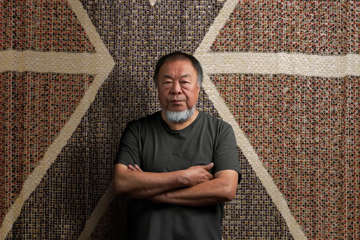 Çağdaş sanatçı Ai Weiwei, Manchester'daki Button Up! sergisi kapsamında milyonlarca küçük düğmeden oluşan, kırmızı, beyaz, mor ve kahverengi tonlarında geometrik desenler barındıran devasa bir enstalasyonun önünde kolları bağlı ve düşünceli bir ifadeyle duruyor.
