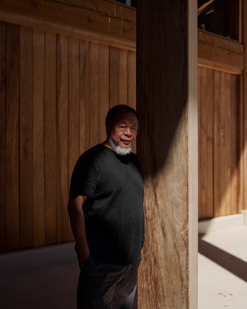 Koyu renk tişört ve pantolon giymiş, gri sakallı sanatçı Ai Weiwei, yüzünün sağ tarafına düşen güçlü güneş ışığıyla belirginleşirken, arkasındaki ahşap panel duvar ve kalın ahşap sütun önünde durarak Manchester'daki Button Up sergisinin gözetim, iktidar ve direniş temalarını derinlemesine düşündürüyor.