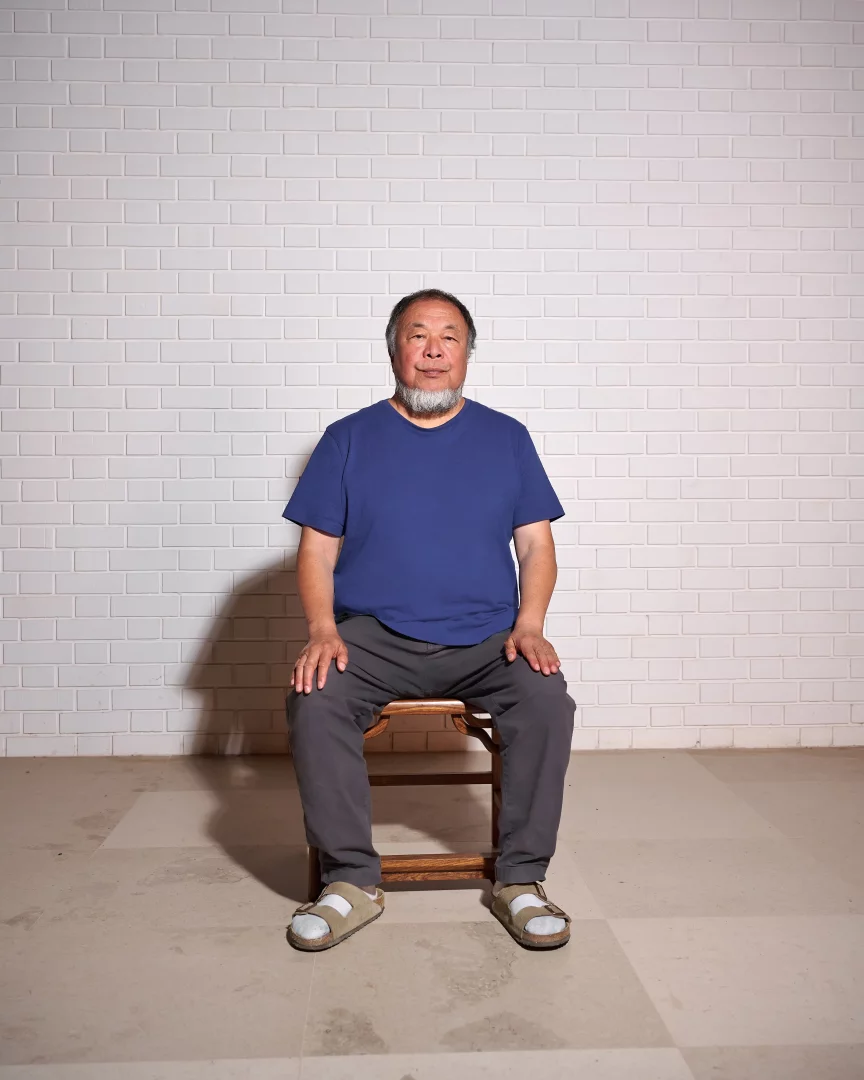 Çinli çağdaş sanatçı ve aktivist Ai Weiwei, Manchester'daki Button Up! sergisi çerçevesinde gözaltı sürecini yeniden canlandıran ve gözetim temasını işleyen performanslarını yansıtan sade bir ortamda, beyaz fayanslı bir duvarın ve açık renkli bir zeminin önünde, gri pantolon ve koyu mavi tişörtüyle bir taburede sakince oturuyor.