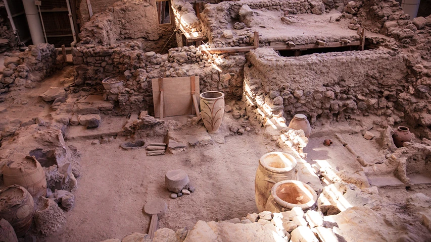 Güneş ışıklarıyla aydınlanan Santorini'deki Akrotiri antik kenti kazı alanında, toprağın katmanları arasından çıkarılmış eski taş duvarlar, düzensiz toprak zemin üzerinde duran çeşitli boyutlarda seramik kaplar ve pithoslar, antik bir yerleşime ait kalıntıları gözler önüne seriyor.