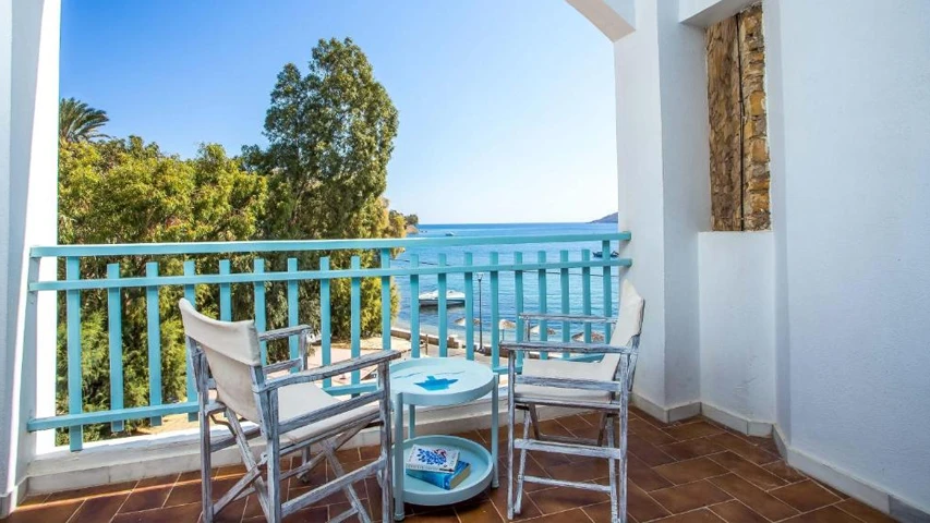 Leros'taki Alea Mare Otel'in deniz manzaralı balkonunda, açık mavi korkulukların ardında sakin turkuaz denizi ve ağaçlık kıyıyı gören, iki beyaz ahşap sandalye ve ortalarında küçük mavi bir sehpa ile rahat bir oturma alanı bulunmaktadır.