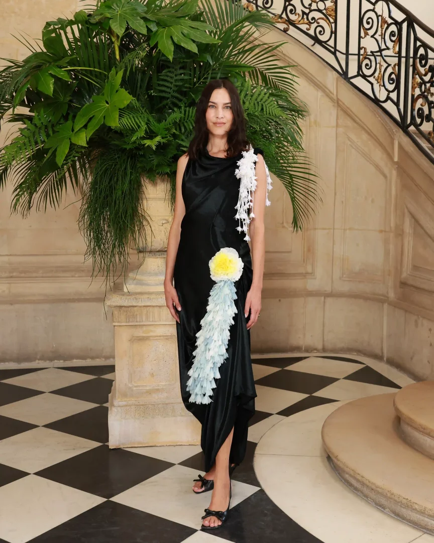 Alexa Chung, Paris Haute Couture Moda Haftası'nda Dior'un sanatsal couture yaklaşımını yansıtan, dökümlü siyah elbisesi üzerindeki heykelsi beyaz, sarı ve açık mavi çiçeksi ve tüylü işlemeleriyle şık bir duruş sergiliyor.