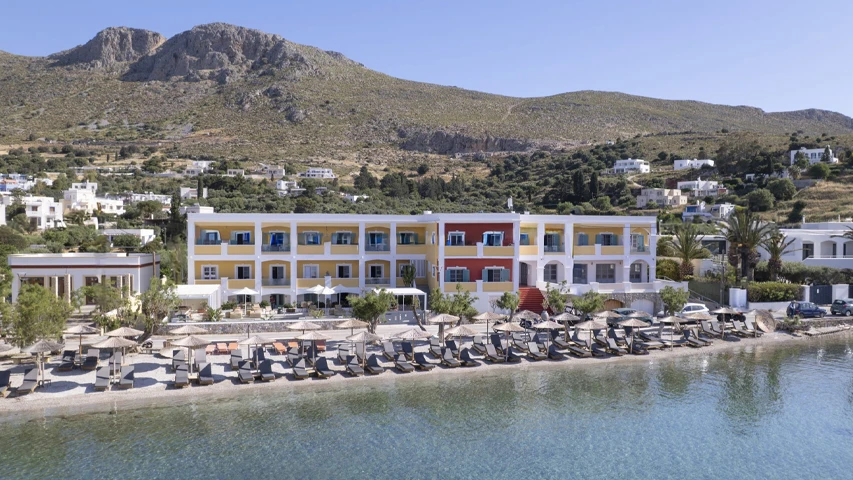 Leros'taki Alidian Bay Suites, sarı ve kırmızı cepheli binasının önündeki plajda dizili şezlonglar ve hasır şemsiyelerle, berrak denizi ve arka plandaki kayalık tepelerde yer alan beyaz evlerle sıcak, güneşli bir günün keyfini sunuyor.