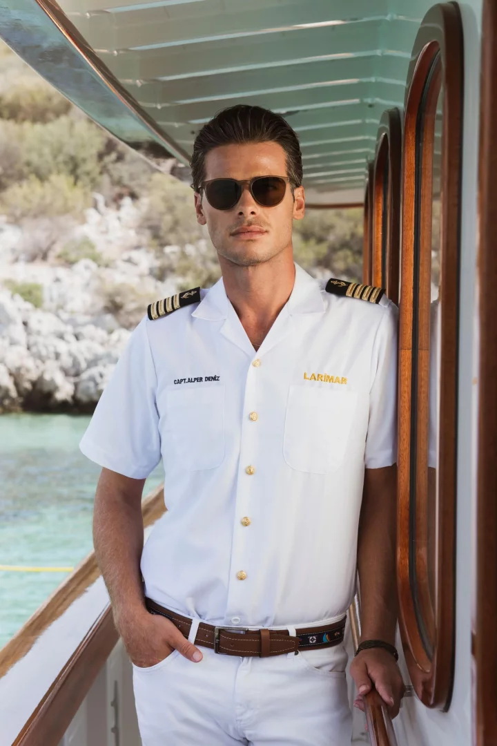 Filo Kaptanı Alper Deniz, omuzlarında rütbe işaretleri olan beyaz üniformasıyla ve güneş gözlükleriyle LARİMAR yatının güvertesinde, berrak turkuaz sulara nazır, kayalık bir kıyının önünde mavi yolculuğun keyfini çıkarırken karizmatik bir şekilde poz veriyor.