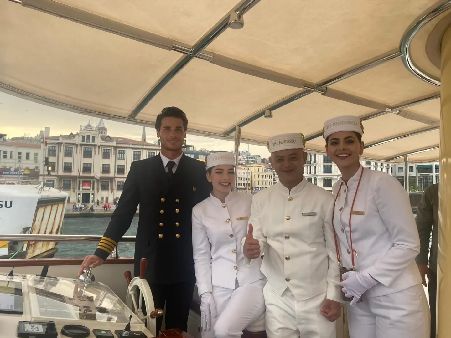 Filo Kaptanı Alper Deniz, kaptan üniforması içinde teknenin kontrol panelinde dururken, Peninsula otelinin beyaz üniformalı güler yüzlü üç ekibiyle birlikte, Galata Kulesi'nin belirgin olduğu İstanbul denizinde keyifli bir mavi yolculuk deneyimini yansıtıyor.