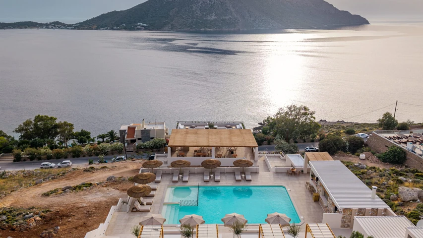Kalimnos'taki Analeia Luxury Hotel'in kuşbakışı görüntüsünde, masmavi denizin ve dağların huzurlu manzarasına karşı konumlanmış, güneşlenme şezlongları ve hasır şemsiyelerle çevrili pırıl pırıl bir yüzme havuzu, zarif restoran ve modern yapılar belirgin şekilde görülüyor.