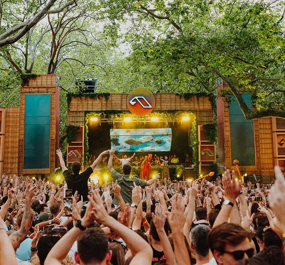 Anjunadeep Open Air etkinliğinde, ağaçlarla çevrili bir orman atmosferinde, ahşap ve bitkilerle süslenmiş büyük bir sahnede performans sergileyen DJ'lerin önünde, yüzlerce elektronik müzik tutkunu ellerini coşkuyla havaya kaldırarak bu açık hava deneyiminin tadını çıkarıyor.