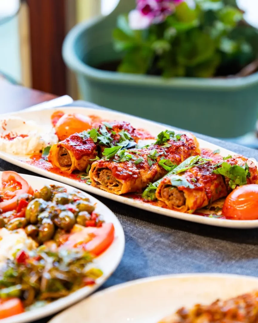 Büyük beyaz bir tabakta kırmızı soslu ve taze yeşilliklerle süslenmiş rulo et yemekleri, yanında zeytin salatası ve domates dilimleri bulunan diğer bir tabak ile Antakya mutfağının lezzetli çeşitliliğini sergiliyor, arka planda yeşil bir saksı bitkisi seçiliyor.