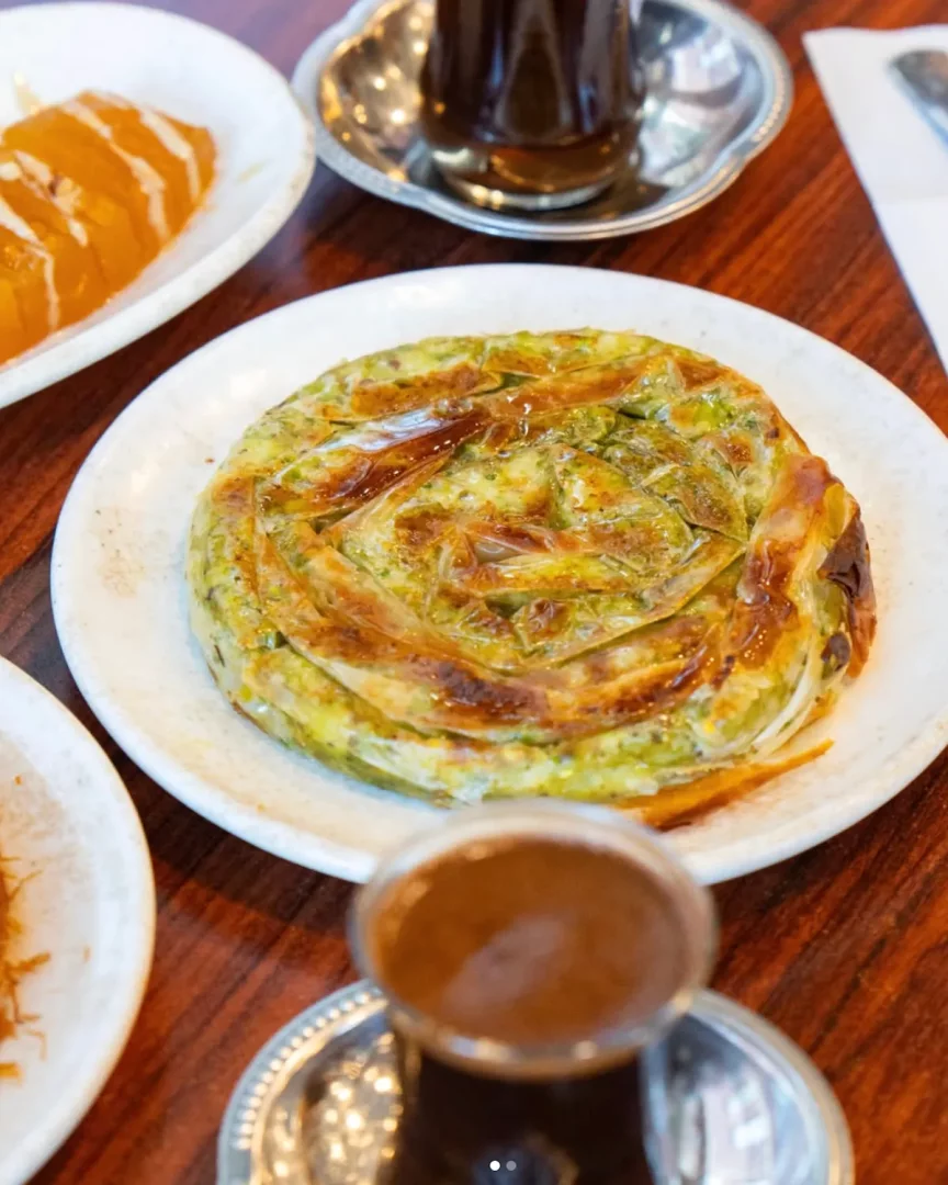 Kahverengi ahşap bir masa üzerinde, Antakya mutfağının geleneksel lezzetlerinden biri olan, altın rengi katmanlı ve yeşil otlu dolgulu yuvarlak bir börek tabağı, arka planda çay ve sol yanda dilimlenmiş turuncu renkli bir tatlı ile sunuluyor.